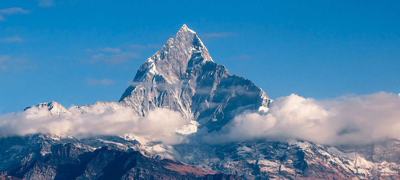 A sharp, snow-capped mountain peak rises above clouds under a blue sky. A sharp, snow-capped mountain peak rises above clouds under a blue sky.