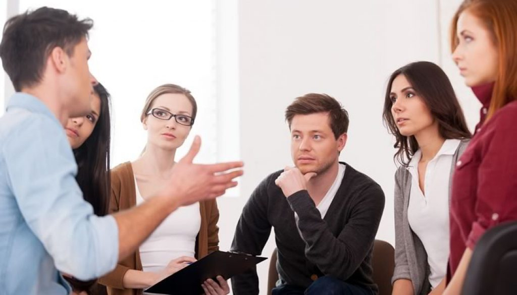 A group in a therapy session listens intently to a speaker's hand gesture. A group in a therapy session listens intently to a speaker's hand gesture.