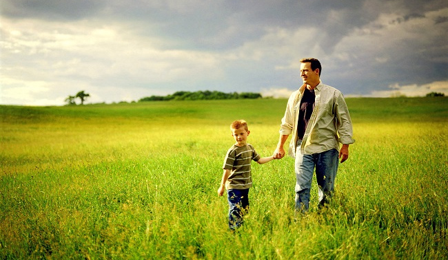 A father and son walk hand in hand through a green field under a cloudy sky.