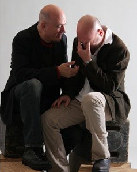 Two men sitting closely, one comforting the other who appears distressed. Two men sitting closely, one comforting the other who appears distressed.